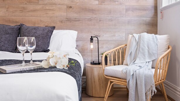 A neatly arranged bedroom with a bed, two pillows, a bedside table with a lamp, a wicker chair, and a book. - Hidden Cozy Gem In Caulfield North (Caulfield North)