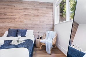 A hotel room with a wooden wall, a bed with white bedding, a wicker chair, a bedside table with a lamp, and a large mirror. - Hidden Cozy Gem In Caulfield North (Caulfield North)