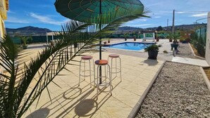Ein Poolbereich mit einem großen Sonnenschirm, drei Barhocker und Blick auf Berge im Hintergrund.