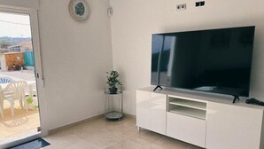Ein Wohnzimmer mit einem großen Flachbildfernseher auf einer weißen Unterhaltungseinheit, einer kleinen Topfpflanze und einer Tür, die zu einem Außenbereich mit Stühlen und einem Tisch führt.