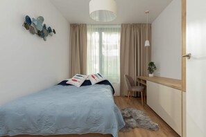 A bedroom with a bed, a desk, a chair, a plant, and a window with curtains. - Mielno | 11 Piastów Street | Modern Apartment | Sea View (Mielno)