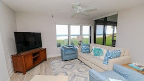 Sala de estar com televisão, sofás e uma grande janela com vista para a praia.