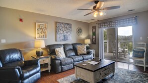 A living room with a leather sofa, coffee table, and a ceiling fan. There is a balcony with chairs and a view of the outdoors. - FREE Dollywood Ticket • Whispering Pines 143 • 2bd/2ba • City View • Pools (Pigeon Forge)