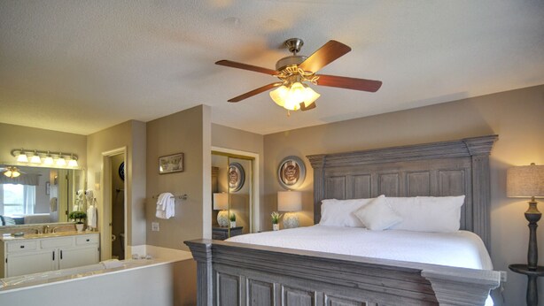 A bedroom with a large bed, a ceiling fan, a nightstand, a lamp, and a bathroom with a bathtub and vanity. - FREE Dollywood Ticket • Whispering Pines 143 • 2bd/2ba • City View • Pools (Pigeon Forge)