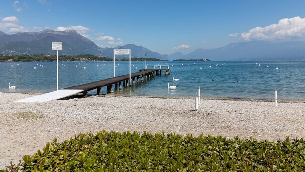 A pebbled beach with a wooden pier extending into calm waters, surrounded by mountains and clear skies. - Il Professore: front lake apartment (Manerba del Garda)