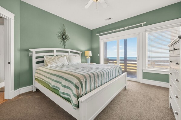 A bedroom with a bed, a nightstand, a lamp, a dresser, and a view of the ocean through a sliding glass door.