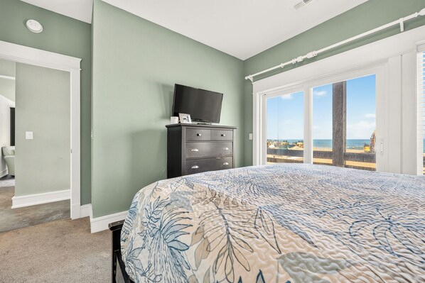 A bedroom with a bed, a dresser, a television, and a large window with a view of the beach.