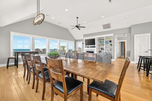 A spacious dining area with a large wooden table and chairs, a ceiling fan, and a view of the ocean through large windows.