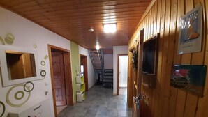 Un couloir aux murs en bois et au sol carrelé, qui mène à un escalier. Il y a des portes sur la droite, dont une avec un téléviseur et une autre avec une pancarte.