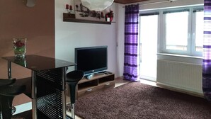 Un salon avec un plafond en bois, un téléviseur posé sur un meuble en bois, une table en verre avec un vase, un rideau violet et une fenêtre avec des stores.