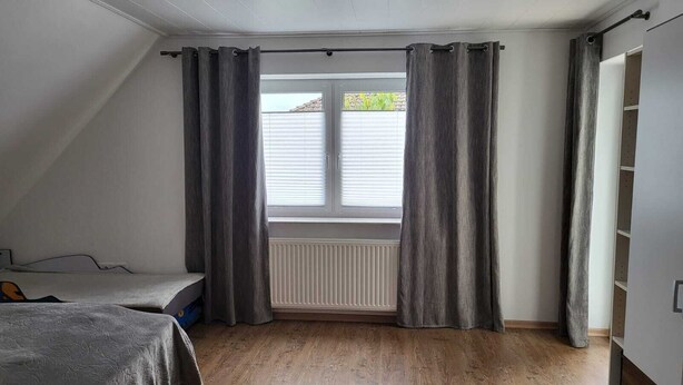 Une chambre avec un plafond en pente, une fenêtre avec des stores blancs, un radiateur placé sous la fenêtre et un lit recouvert d’une couverture grise.