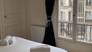Ein Hotelzimmer mit Bett, Heizkörper und Fenster mit Blick auf Gebäude und einen Balkon.