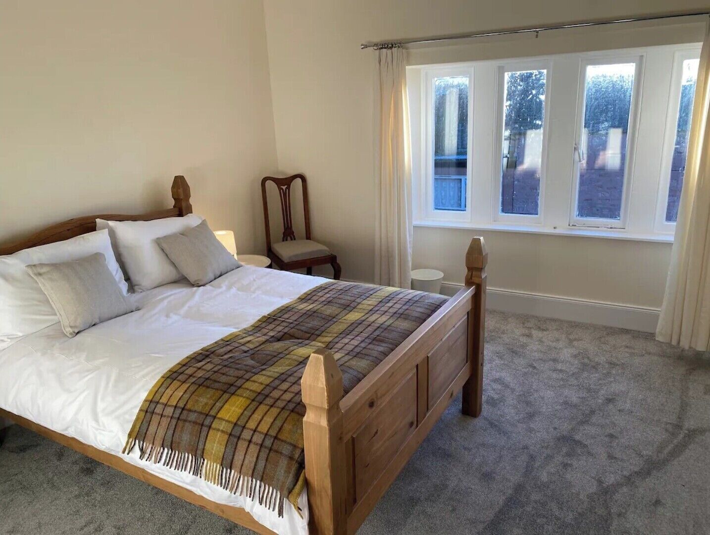 A bedroom with a wooden bed, white bedding, a plaid throw blanket, and a window with curtains.