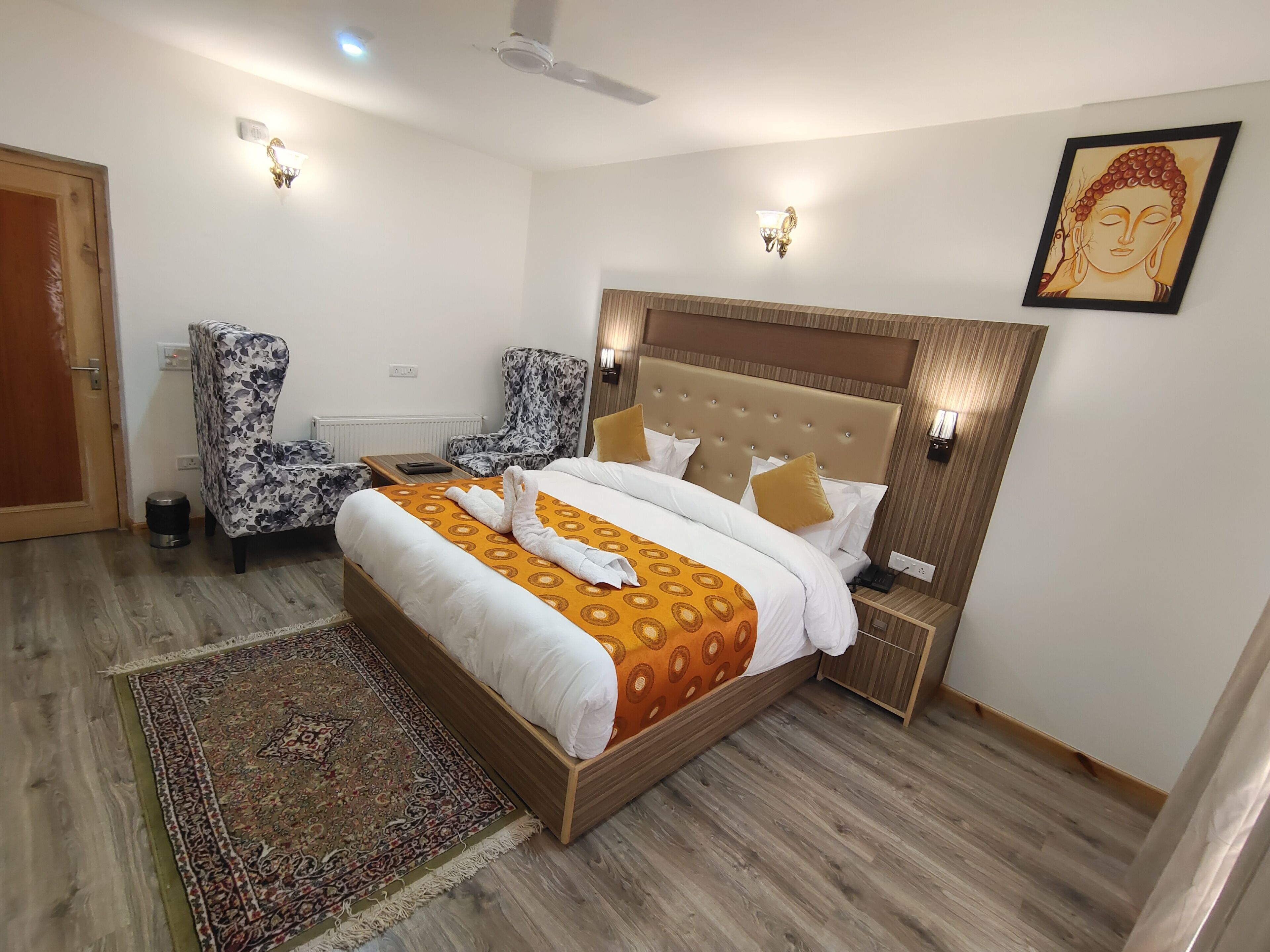 Un cuarto de hotel con una cama grande, dos sillones, una mesita, una mesita de noche, un ventilador de techo y un cuadro de Buda en la pared.
