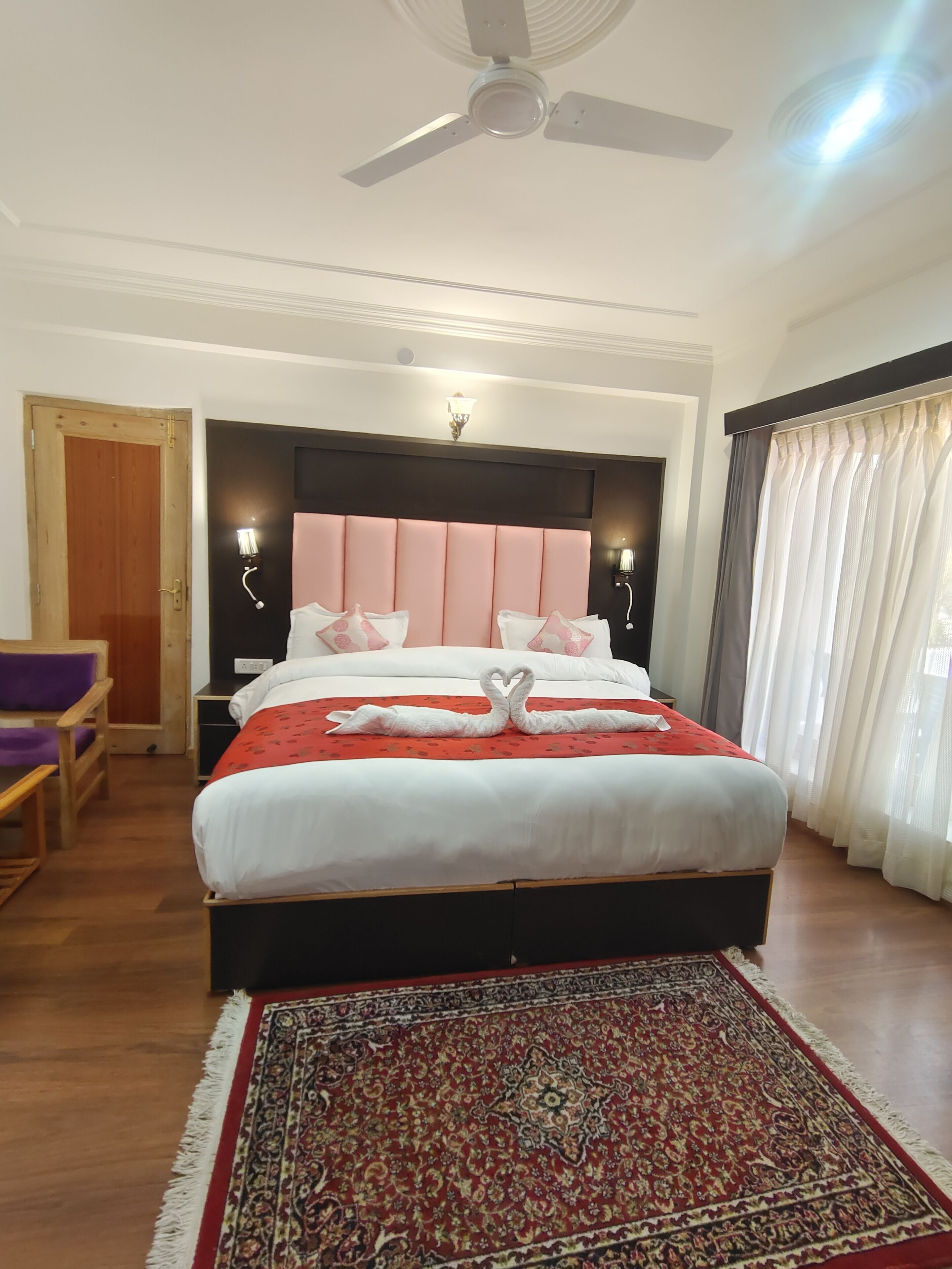 A bedroom with a large bed, a ceiling fan, a wooden door, a small table, and a chair.
