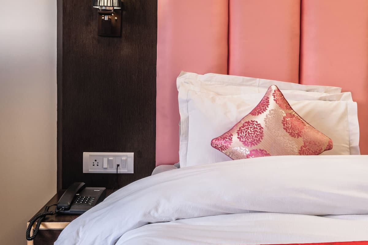 A hotel room with a wooden headboard, a bedside lamp, a telephone, and a bed with white bedding and a red accent.