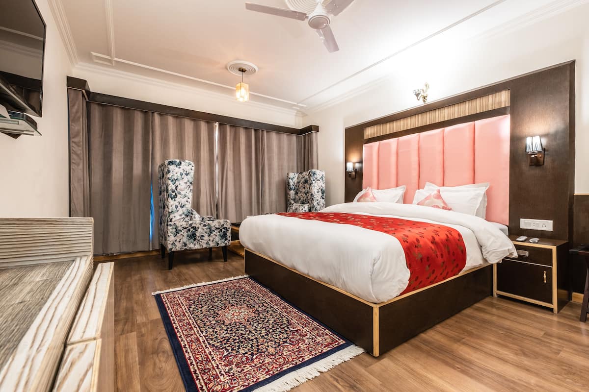 A modern hotel room with a large bed, a sitting area with a sofa and armchairs, a wooden floor, and a ceiling fan.