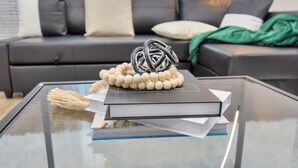 A modern living room with a glass coffee table, a stack of books, and a decorative metal sculpture. - ✵The Allure✵| Houston Hidden Gem - NRG/Med Center (Houston)