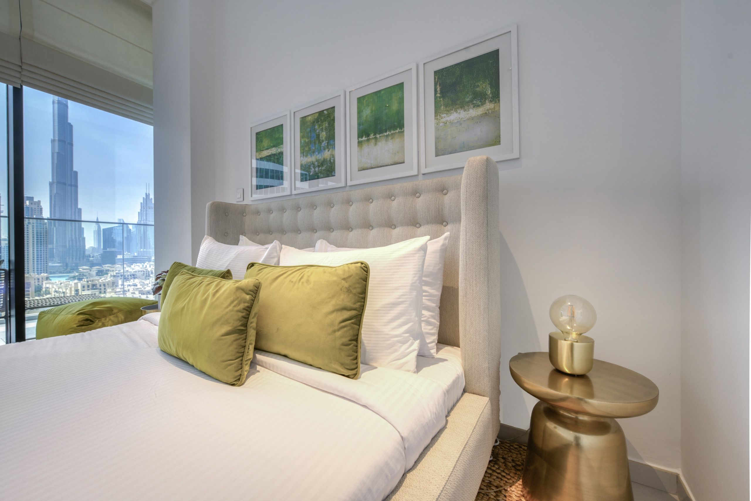 A modern hotel room with a large bed, green and white pillows, a tufted headboard, and framed artwork on the wall.