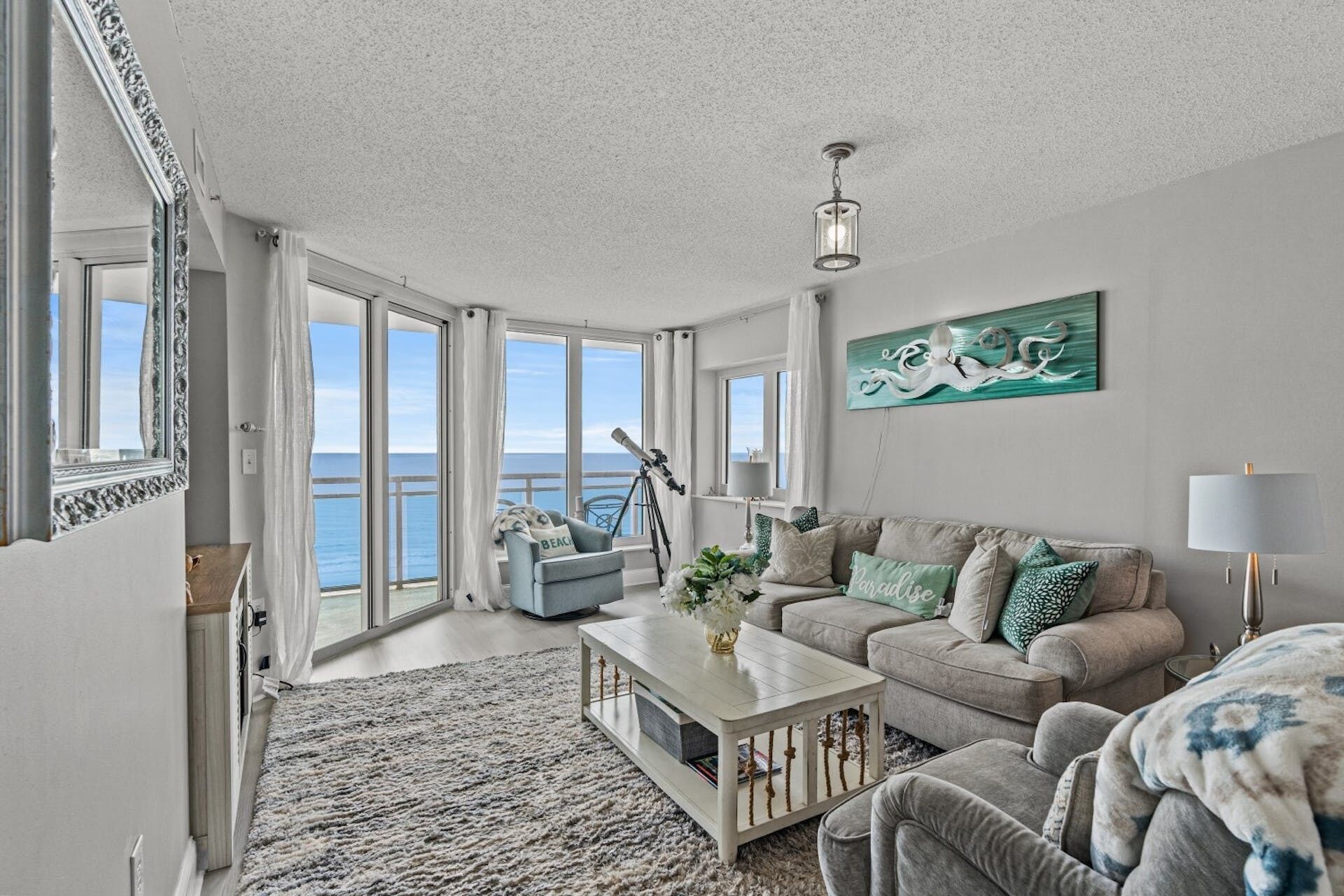 A spacious living room with a large window overlooking the sea, a cozy sofa set, a coffee table, and a telescope.