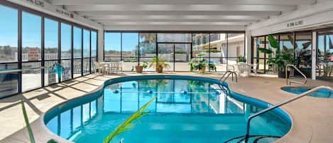 An indoor swimming pool with clear glass walls, a pool table, and several potted plants.