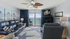 Sala de estar com sofá de couro, poltrona cinza, tapete estampado com estrelas, ventilador de teto e vista para o mar através de uma porta de vidro.