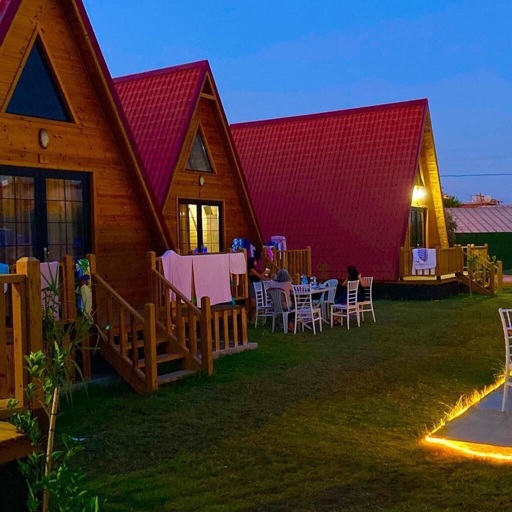 A wooden cabin with a red roof, outdoor seating area with tables and chairs, and a small deck with a railing.