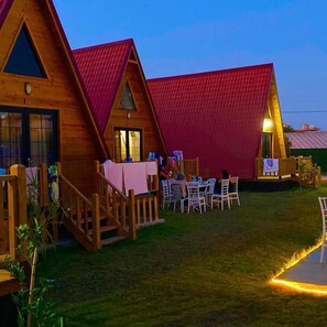 A wooden cabin with a red roof, outdoor seating area with tables and chairs, and a small deck with a railing. - Noel Bungalov Cafe (Demre)