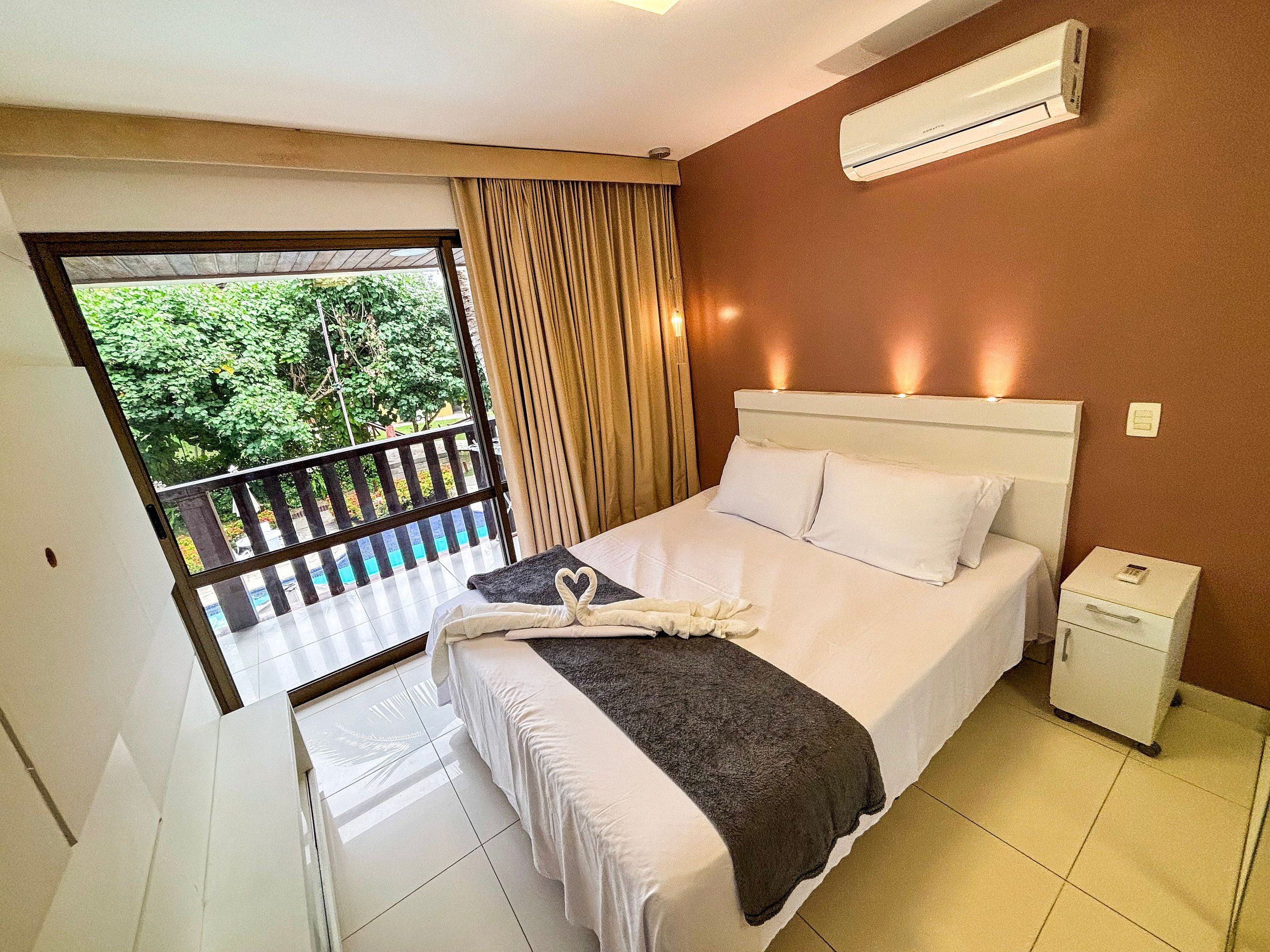 Quarto de hotel com cama, mesa de cabeceira, ar condicionado e varanda com vista para o verde.