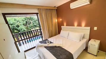 Quarto de hotel com cama, mesa de cabeceira, ar condicionado e varanda com vista para o verde.