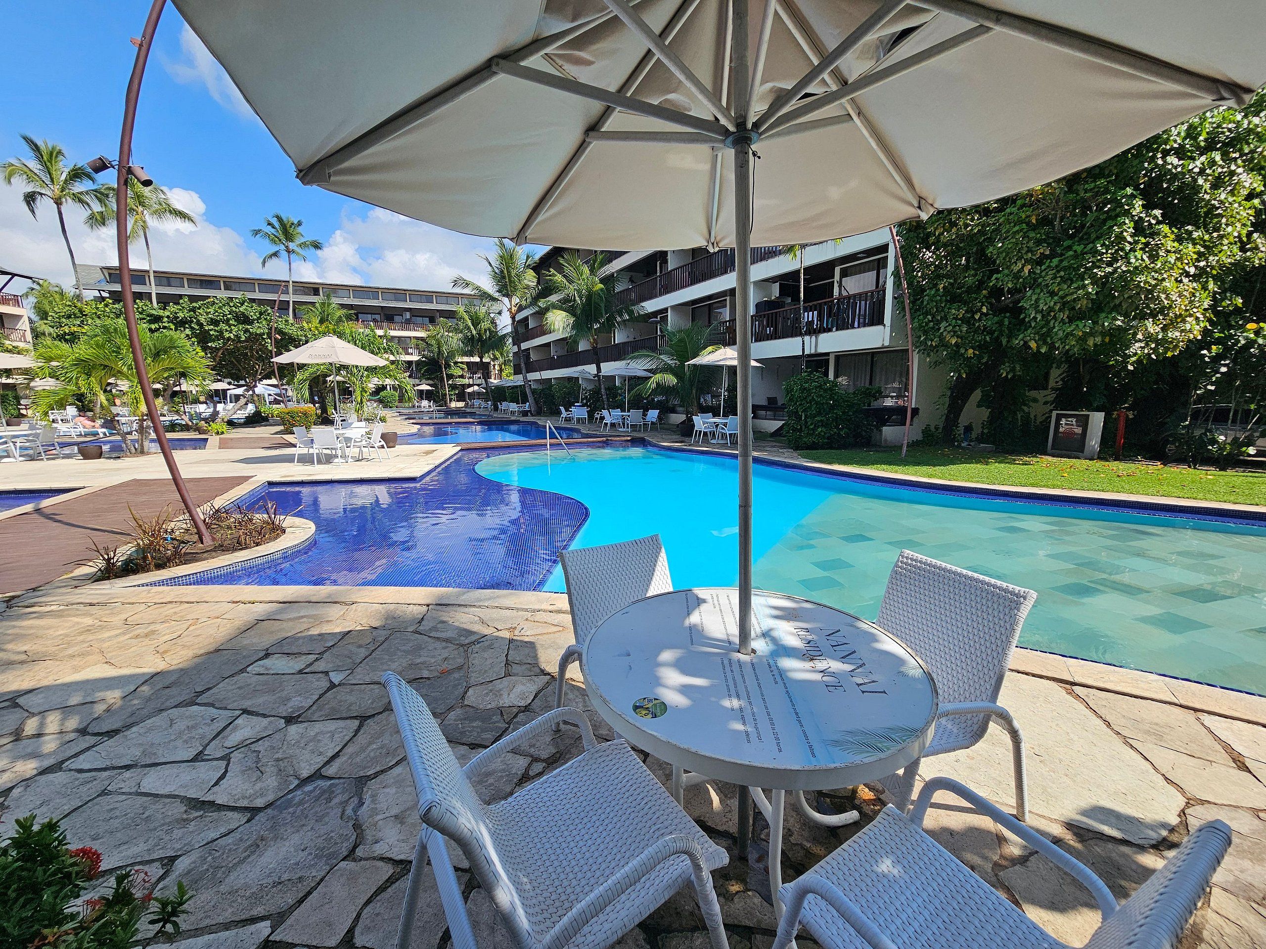 Piscina com espreguiçadeiras e guarda-sóis, cercada por vegetação e um edifício de vários andares.