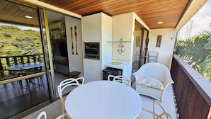A balcony with a table and chairs, a kitchen area with an oven, and a view of greenery. - Nannai Residence (Ipojuca)