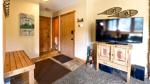 A wooden table, a television, and a door in a room with a carpeted floor and a wall-mounted picture.