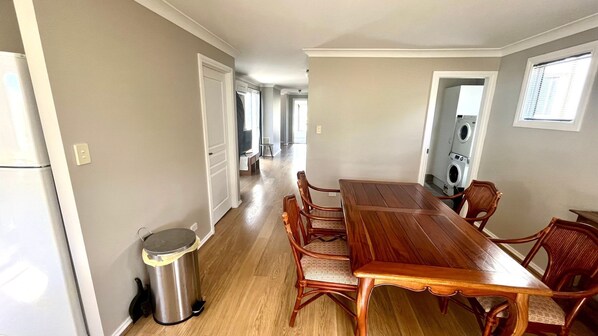 Un coin repas avec une table et des chaises en bois, une poubelle et une buanderie visible à travers une porte ouverte.