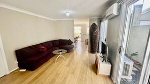 Un salon avec un canapé rouge foncé, une petite table ronde et un plancher en bois. Un grand vase décoratif et un appareil de climatisation blanc se trouvent sur le côté droit.