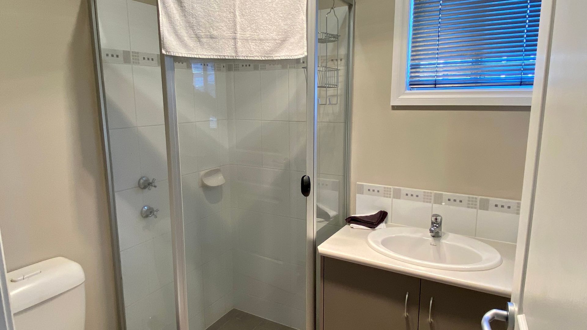 Un baño con cabina de ducha de vidrio, un lavabo blanco y una ventana con persianas.