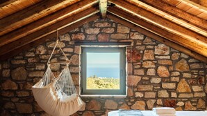 Ein gemütliches Zimmer mit Steinwand, einer Hängematte, einem Bett mit Handtüchern und Blick auf das Meer durch ein Fenster.
