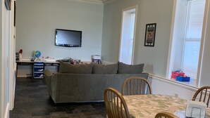 A cozy living room with a gray sofa, wooden chairs, a dining table with a leaf-patterned tablecloth, a TV on the wall, and a window with blinds. - One-of-a-kind! Former Schoolhouse turned mansion for rent! The entire place!! (Port Austin)
