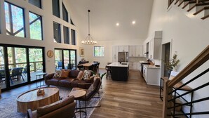 Amplia sala de estar con un sofá de cuero, una mesa de centro de madera y una mesa de comedor. Se observa una cocina con gabinetes blancos y un refrigerador de acero inoxidable. A la derecha se ve una escalera con barandillas de madera.