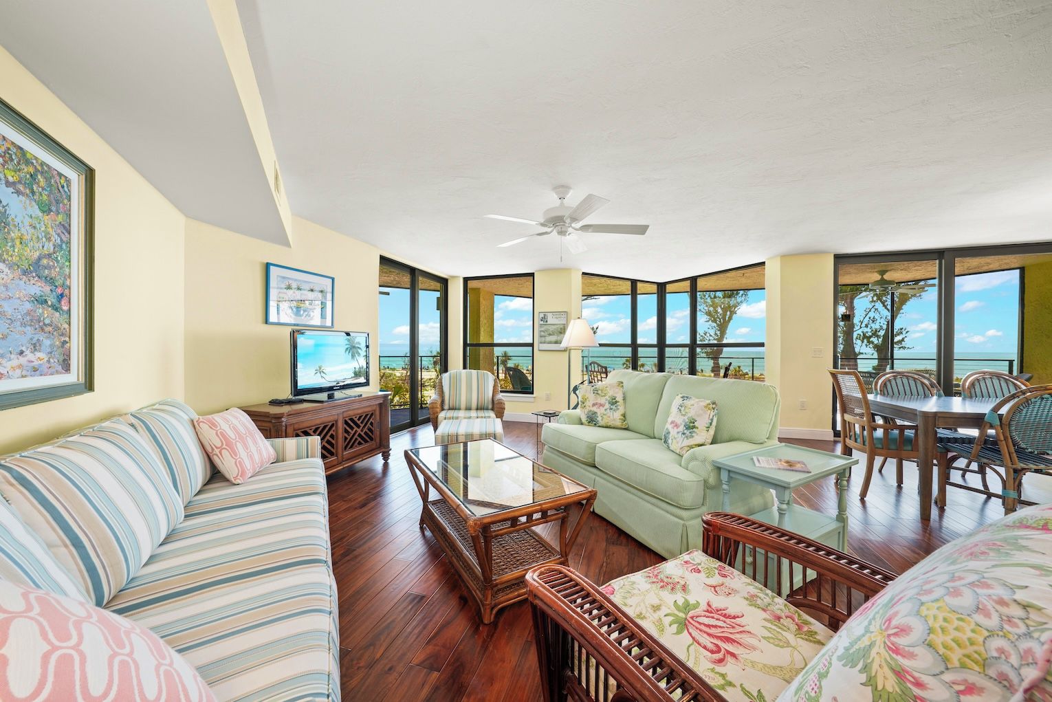 A spacious living room with a large window offering a view of the ocean, a ceiling fan, and a television on a wooden cabinet.