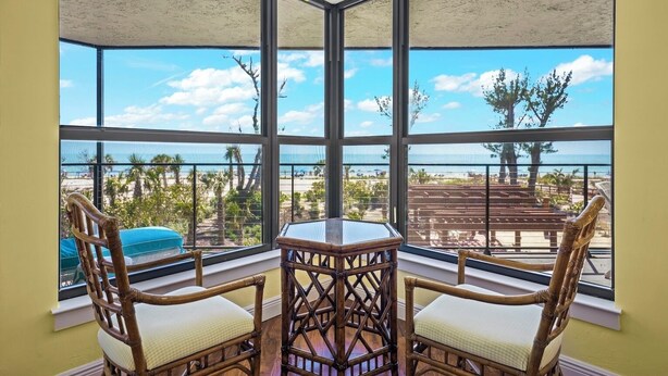 Habitación de esquina con ventanas amplias que dan a una playa, con sillas de mimbre y una mesita.