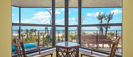 Habitación de esquina con ventanas amplias que dan a una playa, con sillas de mimbre y una mesita.