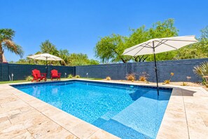 A rectangular swimming pool with a blue tile edge, surrounded by a stone deck. There are two red Adirondack chairs and a white umbrella.