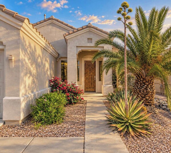 A residential building with a front entrance, a pathway, and various plants including a palm tree and flowering shrubs.