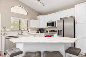 A modern kitchen with white cabinets, a large island, stainless steel appliances, and a window with blinds.