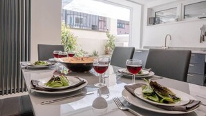 Mesa de jantar posta com pratos de comida e copos de vinho tinto numa cozinha moderna com vista para edifícios.