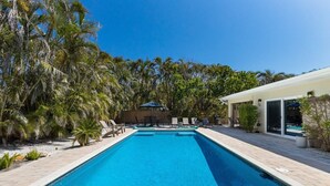 A rectangular swimming pool with lounge chairs and umbrellas, surrounded by palm trees and a covered patio area. - The Salted Coconut 3 Bedroom Home by RedAwning (Anna Maria)