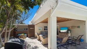 An outdoor patio area with a barbecue grill, wicker furniture, and a TV screen displaying a beach scene. - The Salted Coconut 3 Bedroom Home by RedAwning (Anna Maria)