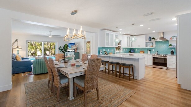 A modern kitchen with white cabinets, a central island, and a dining area with wicker chairs. - The Salted Coconut 3 Bedroom Home by RedAwning (Anna Maria)