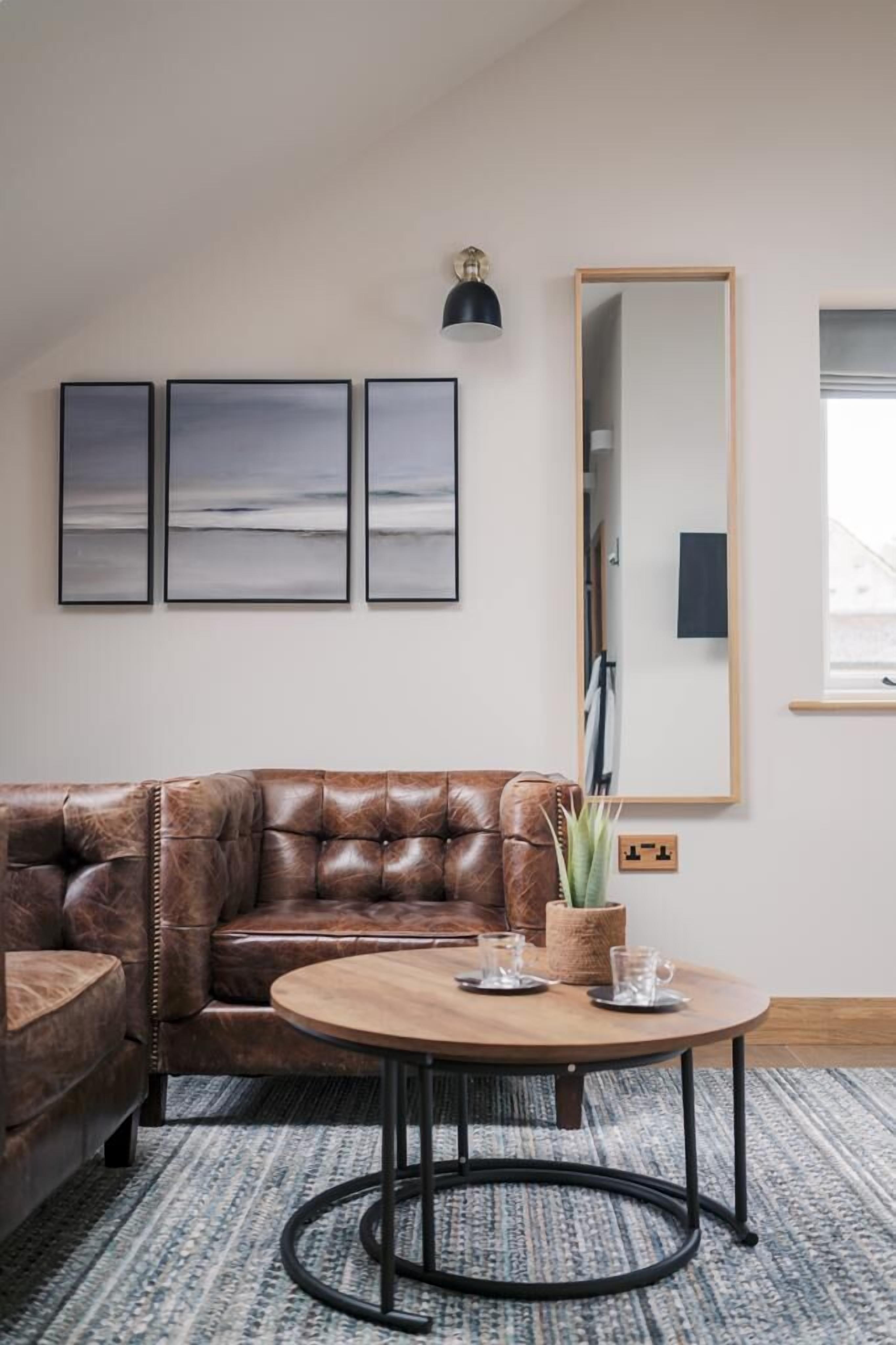 A living room with a brown leather sofa, a round wooden coffee table, and three framed artworks on the wall.